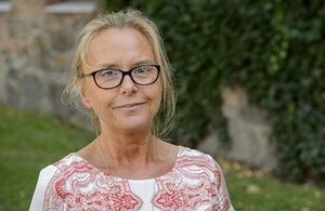 Lund University Director Susanne Kristensson is photographed outdoors. She has blond hair styled up in a bun and dark glasses. She is smiling and looking at the camera. She is wearing a short-sleeved white blouse with a red paisley pattern. 