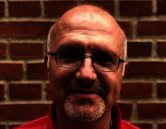 Jordan stands in front of a red brick wall looking straight at the camera. He is wearing black steel-rimmed glasses and a red polo shirt. He has short hair, a short beard and a moustache.