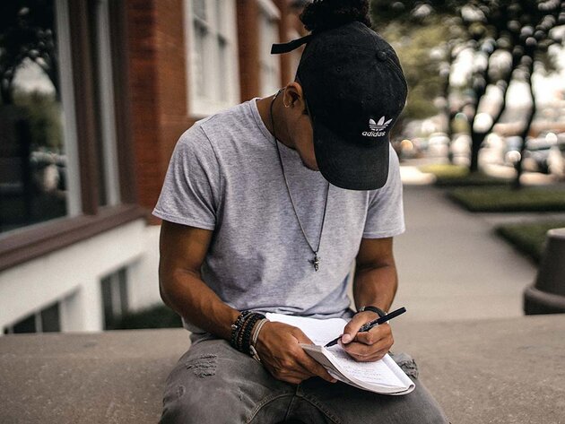 A young man is sitting on a stone ledge writing in a pad. He is looking down at the text and wearing a black Adidas cap, so you cannot see his face. He is wearing a grey t-shirt and black jeans with patches. He has several bracelets around his right wrist and a silver pendant around his neck.