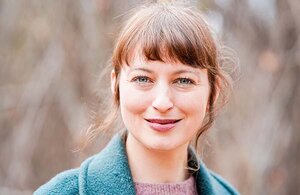 Katja is smiling and looking at the camera. She has blue eyes and red hair put up in a bun. Strands of hair hang down in front of her ears. She is wearing a pink knit sweater and a light blue coat. She is standing outdoors, with the wind blowing her hair slightly to the side.