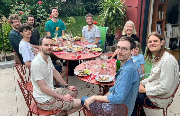 On an outdoor veranda around two tables, a group of nine people are sitting and looking happily at the camera. Jakob is sitting in the lower right corner of the picture, wearing a light-coloured button-down shirt and dark jeans. He is smiling.  