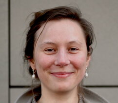 Katarzyna is smiling broadly and looking at the camera. She has brown hair styled up in a bun and pearl earrings. She is wearing a small-checked shirt and a grey coat over it.  