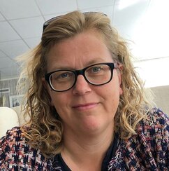 Elisabet is smiling and looking into the camera. She has short, light curly hair held back by sunglasses on her head. She has blue eyes and black-framed glasses. She is wearing a black and white patterned blouse.