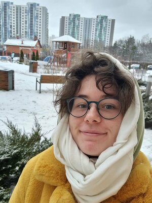 Yevheniia is standing outside, in front of a playground. It is winter and she has a woollen scarf tied around her head. She has dark curly hair and glasses.