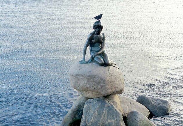 Sitting on a pile of stones at the water's edge is a bronze statue of a little mermaid. It is Edvard Eriksen's famous statue in Copenhagen, “The Little Mermaid”. A bird is resting on the statue's head.