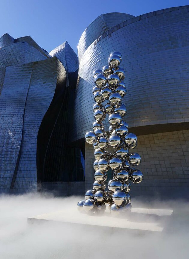 En futuristisk byggnad som är slät och formad i olika kurviga delar är belyst av solen. Nertill i bilden syns vit rök som omger podiet till en skulptur som står framför byggnaden; skulpturen består av mängder av silvriga klot staplade på varandra i en hög formation. Bilden är tagen utanför Guggenheimmuseet i Bilbao, Spanien.