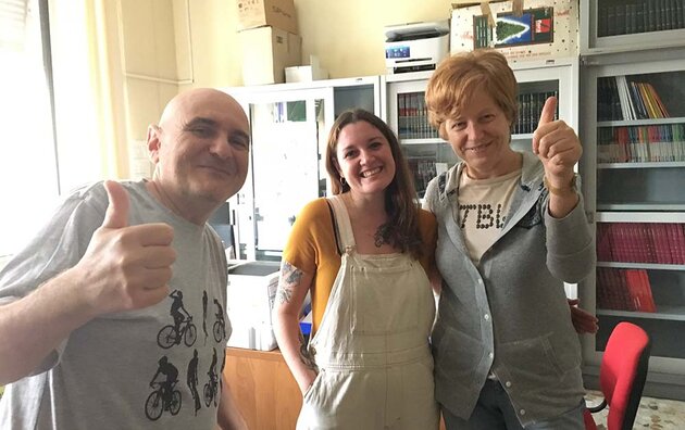 Three smiling people are standing in an office with bookshelves, cabinets and desks. In the centre is Valeria, with long dark hair, a yellow shirt and light-coloured dungarees. She is flanked by two colleagues, both giving her a thumbs up. Fabio Guiffré on the left has a shaved head and a grey shirt with a bicycle motif, while Claudia Magnoni on the right has reddish short hair, a grey cardigan and light-coloured jeans.