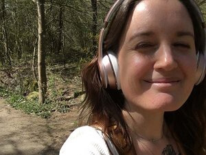 Valeria stands smiling and squinting in the sunshine. She has long dark hair and is wearing silver headphones. The photo is a selfie. Valeria is wearing a white short-sleeved shirt and yellow dungarees. A tattoo can be seen on her right arm, which is extended.