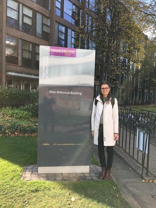 Maria Småberg is standing on campus next to a sign saying “Ellen Wilkinson Building”. She is wearing black trousers and a white coat and appears to have a black backpack on her back. She has long dark hair that is put up with clips. She is wearing glasses. The photo was taken on the University of Manchester campus.