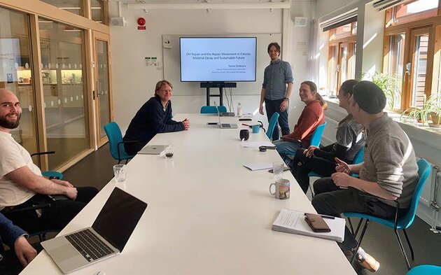 A seminar room with several participants - and Tenno at the head of the table right beside a Power point presentation of his research.