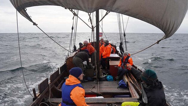 I en öppen träbåt med stort segel ses nio personer som var och en är aktiv på sitt sätt. I förgrunden, i bildens nedre högra hörn, sitter en person med ryggen mot kameran och pekar ut riktningen. Man är ute på öppet hav, både himmel och hav är grått.