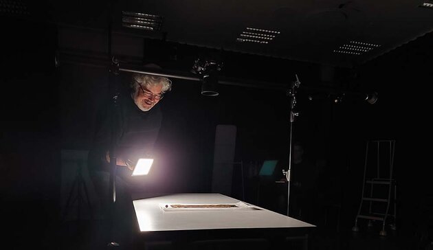 A dark room is illuminated by a single lamp. In the light we see a table with a piece of paper on it, a smiling Stefan Lindgren holding the square light source and a stand above the table with a camera on it.