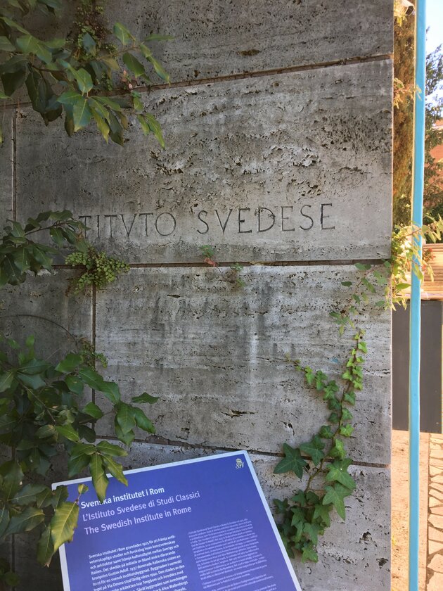 A grey wall has the letters “Istituto svedese” carved in Roman style. Green plants surround the wall from different directions. At the bottom left is a blue metal sign filled with text telling you in three languages that you have arrived at the Swedish Institute in Rome. To the right of the wall is a courtyard.