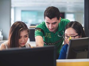 Three young people are facing the camera. Two smiling women with long dark hair are sitting at computers. Behind them is a man with short dark hair, stubble and a green printed shirt. He is leaning forward, pointing at one of the screens. All three of them are looking where he is pointing.