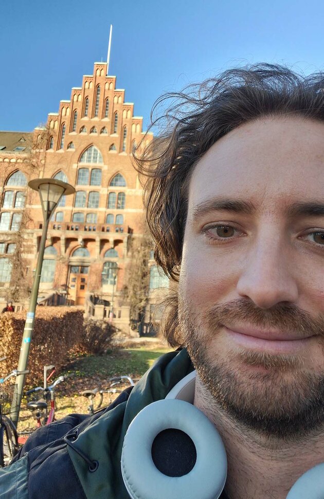 A selfie of Emilio, taken in front of the Main University Library. He has dark hair and beard, and has a pair of head phones around his neck.