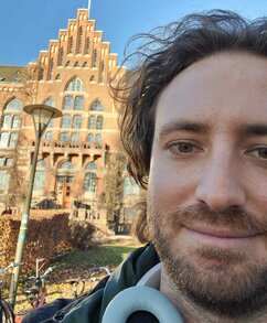 Emilio har tagit en selfie framför Universitetsbiblioteket i Lund. Hans ansikte tar upp den högra halvan av bilden. Han har mörkt lockigt hår, bruna ögon, skägg och mustasch. Emilio ler lite och har ett par hörlurar runt halsen.