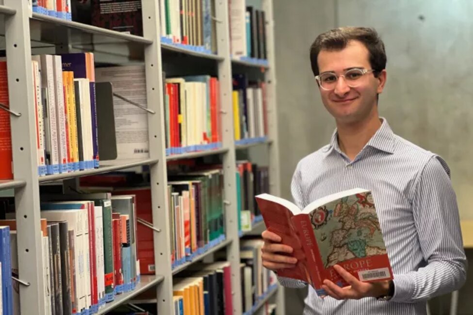 Iveri Kekenadze Gustafsson står m ed en bok uppslagen i handerna bredvis en biblioteksbokhylla och tittar in i kameran.