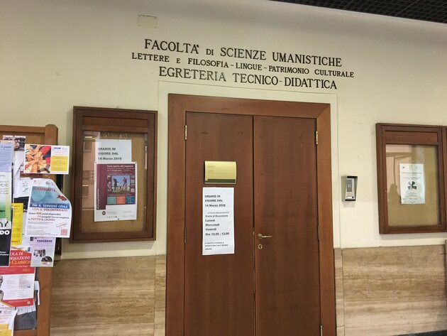 A sign and a posted notice hang on a closed brown wooden door with a brass handle. On either side of the door are notice boards and various posters. Above the door, metal letters embedded in the wall describe which faculty and subjects are hidden behind the door. A letter is missing.