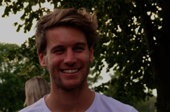 Billy smiles broadly and looks to the side. He has short blond hair and stubble. He is wearing a white T-shirt. The photo was taken outdoors. Greenery and another person, facing away, can be seen in the background.