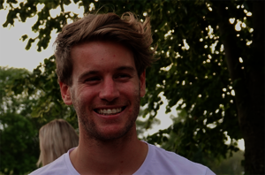 Billy smiles broadly and looks to the side. He has short blond hair and stubble. He is wearing a white T-shirt. The photo was taken outdoors. Greenery and another person, facing away, can be seen in the background.