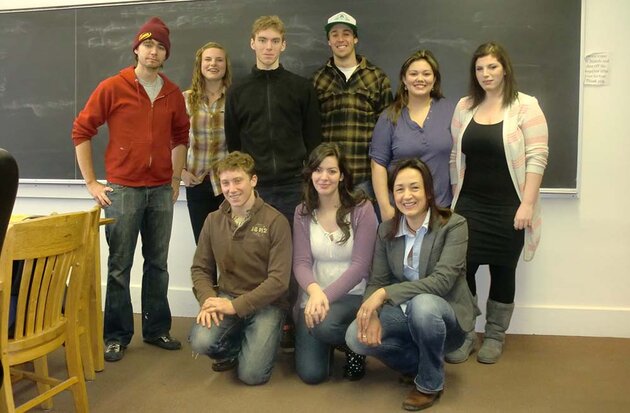 In a lecture hall, in front of the blackboard, nine people pose, six standing and three squatting in front of them. All are young students, except for the person sitting on the bottom right, who is their teacher Blaženka Scheuer from Lund.