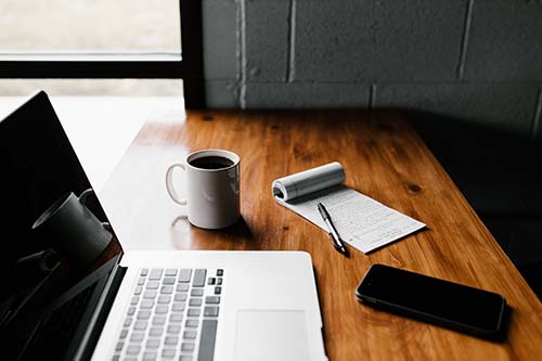 The picture shows a wooden table with several things on it: an open laptop, a mobile phone, a pad and a pen. A white mug filled with coffee is beside the computer. There is light coming in from a window in the background.