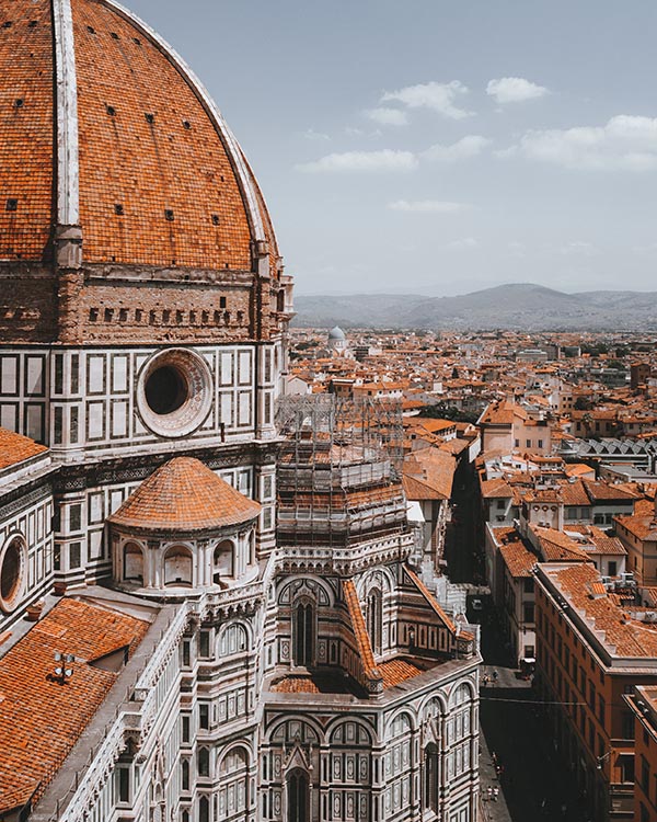 The left part of the picture is occupied by the Cathedral of Santa Maria del Fiore in Florence, with its distinctive architecture and brick-clad dome. The rooftops of Florence and the green hills outside the city are also visible in the distance.
