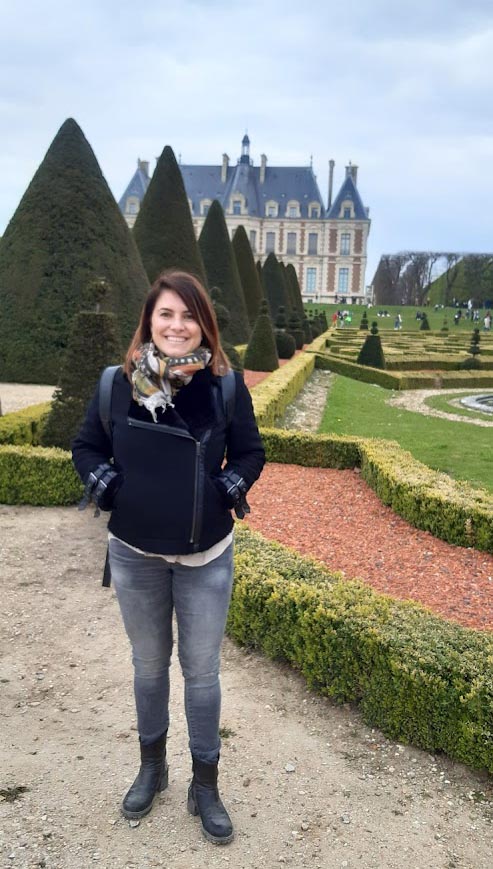 Paola is standing in a formal garden from the 18th century, a beautiful castle in the background. She is in Versailles, outside Paris. She is wearing a black jacket, black jeans and black boots. She wears a colourful scarf around her neck.