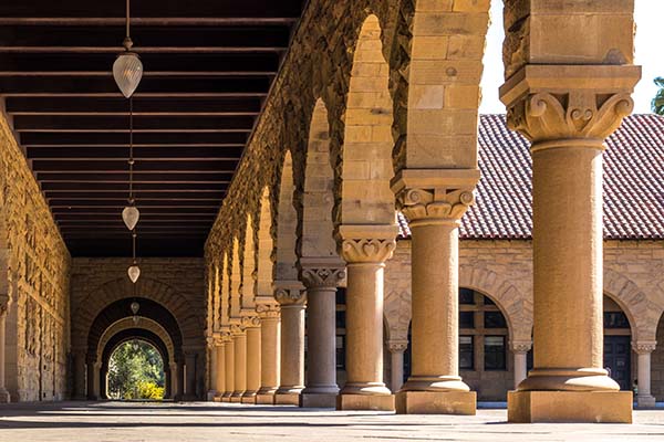 The image is taken inside a column-lined walkway on Stanford University's campus. The sun is coming in from the open space on the right, and the columns are lined up with beautiful arches between them. Drop-shaped glass lamps hang at regular intervals from the ceiling of the columned walkway. At the far end, straight ahead in the picture, the passage opens out into sunlit greenery.