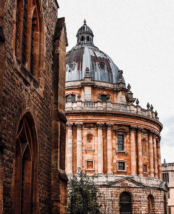 Till vänster i bild syns en vacker röd tegelbyggnad med gotiska fönsterbågar, längre fram tornar den runda 1700-talsbyggnaden Radcliffe Camera upp sig. Bilden är från Oxford.