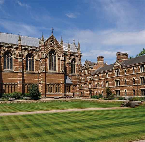 Around a well-mown lawn are ornate red brick buildings. Straight ahead is a church building and to the right lower college buildings. The picture was taken at Keble College in Oxford.