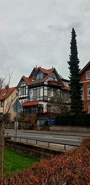 The picture shows a road. On the other side of the road, there is a tall, narrow coniferous tree next to a beautiful white timber frame house with a red tile roof. The picture was taken in Göttingen, Germany.