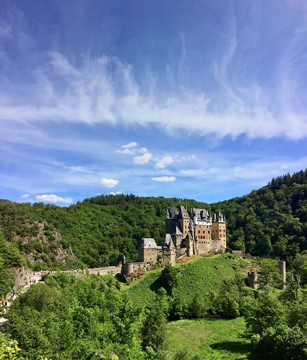 In a green landscape, up on a hill, there is a medieval stone castle with turrets and towers. The wall-lined road leading up to the castle is crowded with tourists. The sky is blue with white clouds that look like wisps of smoke.