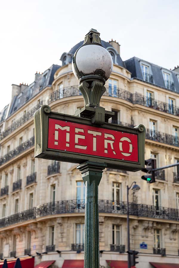 In the foreground of the image is a typical Paris Metro sign, a green metal pole and sign in Art Nouveau style with white text on a red background. In the background, and therefore blurred, is a beautiful building facade.