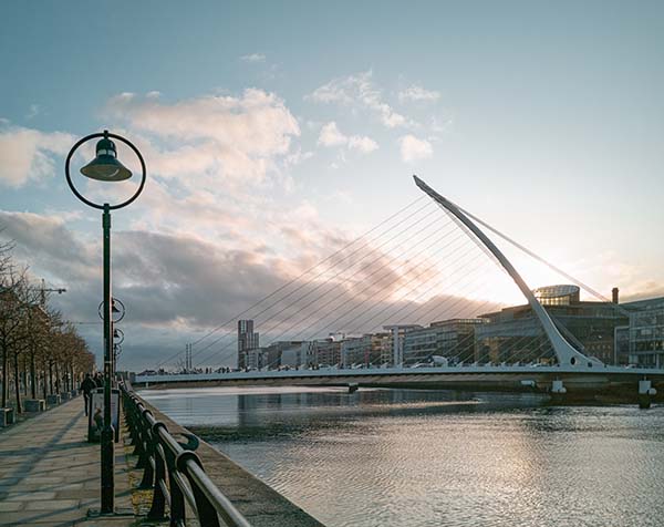 The River Liffey flows calmly. Spanning it is the white, futuristic Samuel Beckett Bridge. There is a pedestrian and cycle path on the left side of the image. The picture was taken early in the morning in Dublin, Ireland.