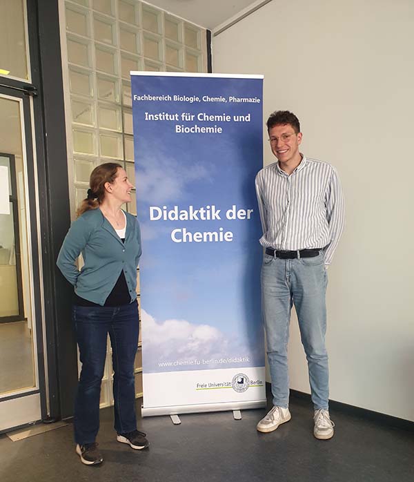 Ylva and her colleague Robert Gieske are standing on either side of a roll-up banner with the text “Didaktik der Chemie”, the didactics of chemistry. Ylva is looking at Robert, while Robert is looking at the camera. Ylva has her hair in a ponytail and is wearing a green cardigan and dark jeans. Robert has short hair and glasses and is wearing a striped button-down shirt and light-coloured jeans.