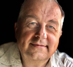 Tomas is seen in close-up so that his face and shoulders fill up almost the whole image. Sunlight falls on his face from the right. He is smiling and looking at the camera. Tomas has short grey hair and is wearing a light grey button-down shirt.